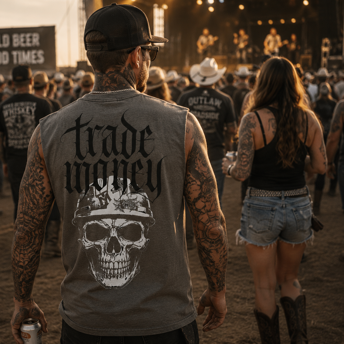 A man standing at a concert or festival, viewed from behind, wearing a gray sleeveless t-shirt with a large white skull and the words 'trade money' printed on the back. He is surrounded by other attendees in a dusty outdoor setting at sunset.